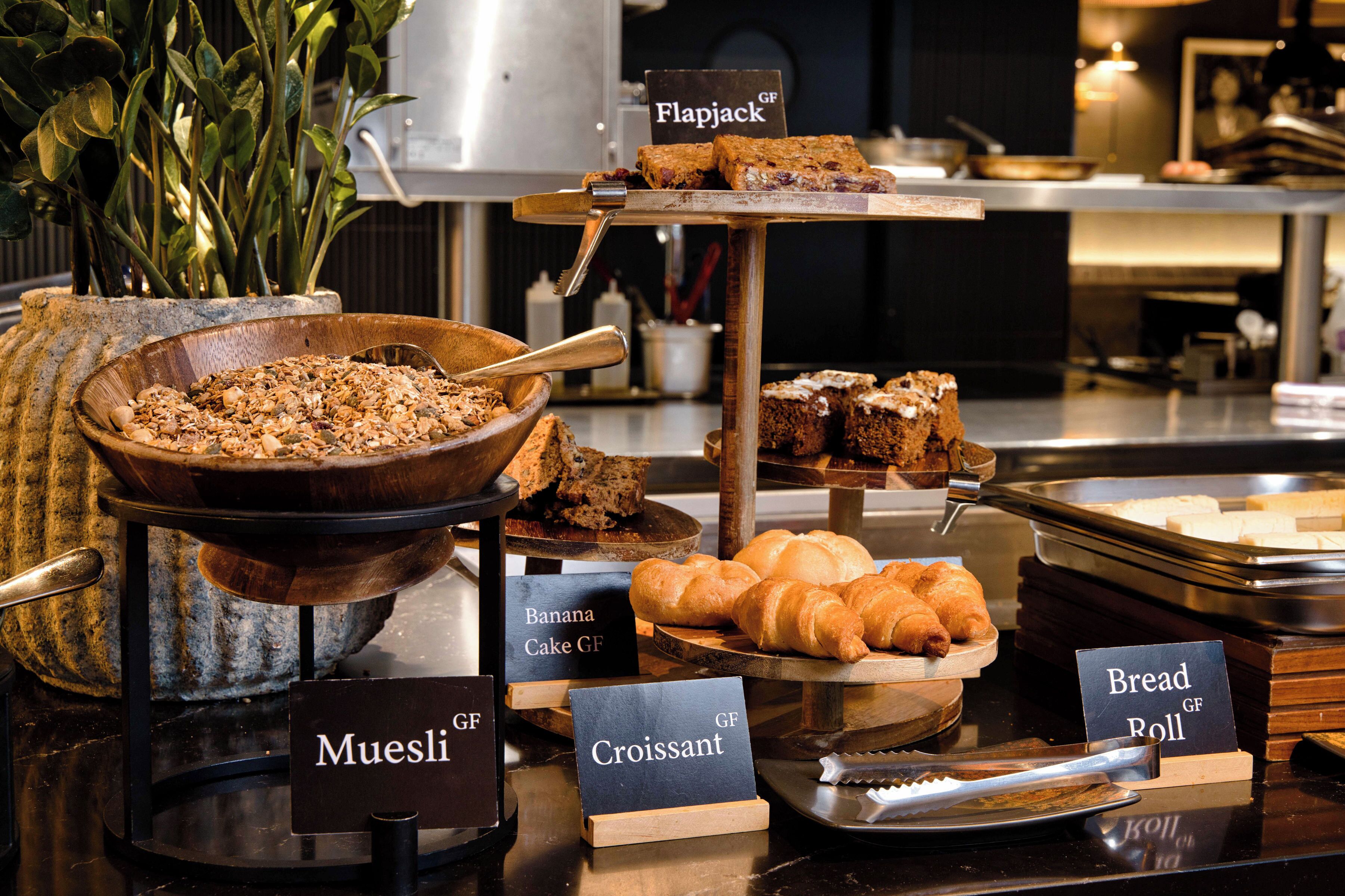 En buffet med tydeligt mærkede retter, herunder mysli, croissanter, pandekager og boller.