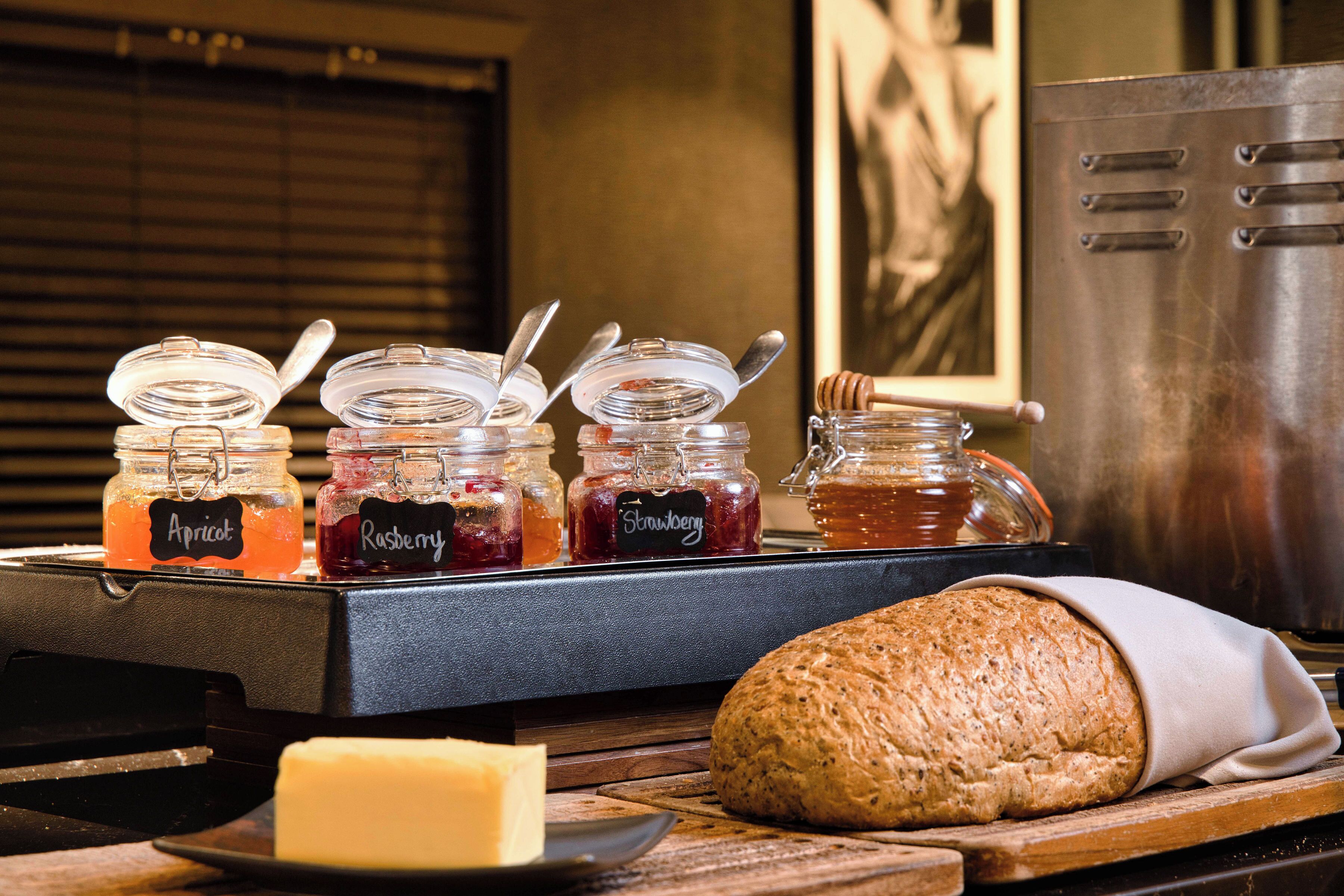A breakfast buffet with jars of jam labeled Apricot, Raspberry, and Strawberry, a loaf of bread, and a plate with a slice of cheese.