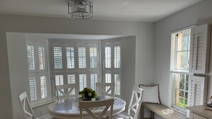 Ein Essbereich mit einem runden Tisch, weißen Stühlen und einer Bank. Große Fenster mit Fensterläden, eine Kristallleuchter und Blick auf Grünanlagen sind zu sehen.