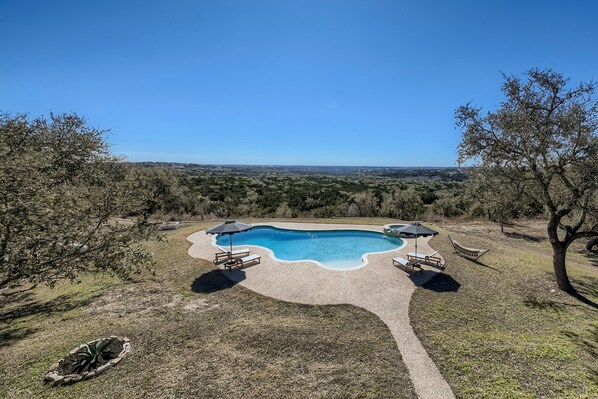 A rectangular swimming pool with a paved deck, surrounded by lounge chairs and umbrellas, set against a backdrop of trees and a distant view. - EcoView Villa•Pool•Spa•Goats•85 acres•Amazing view (Austin)