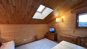 Ein gemütliches Zimmer mit schrägen Wänden, Holztäfelung, einem kleinen Fernseher, einem Schreibtisch und einem Fenster.