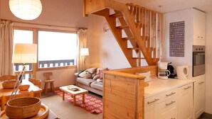 A cozy room with a wooden staircase, a sofa, a dining table with chairs, and a kitchen area with a coffee maker and a kettle.