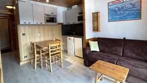 A compact living space with a wooden dining table and chairs, a brown sofa, and a small wooden table. A kitchen area with white cabinets and appliances is visible. - 180m from shops, floor -1, view mountain, balcony, 29m², Méribel-Mottaret (Méribel - Les Allues)