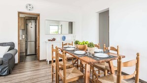Un comedor con una mesa de madera dispuesta para cuatro personas, rodeada de sillas de madera. Se ve una cocina a través de una puerta abierta y un reloj en la pared.