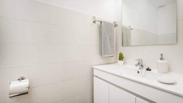 Un baño moderno con azulejos blancos, un espejo grande, un lavabo con grifo, un perchero y un dispensador de papel higiénico.