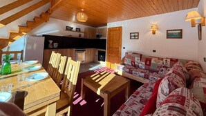 A cozy interior with a wooden staircase, a dining area with a table set for two, a sofa with patterned cushions, and a kitchen in the background. - 180m from shops, floor -1, view mountain, balcony, 60m², Méribel-Mottaret (Méribel - Les Allues)