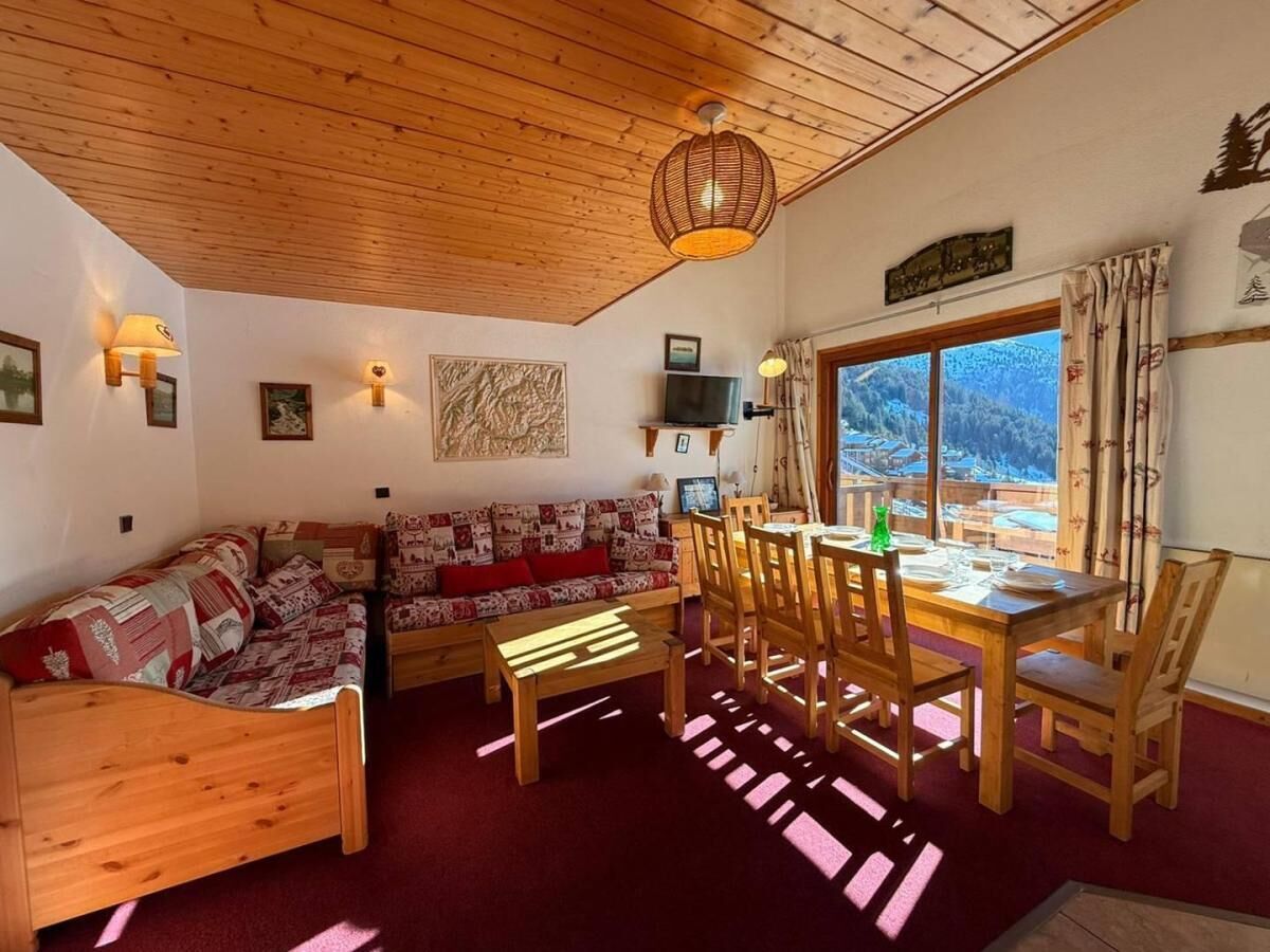 Ein gemütlicher Raum mit Holzbalkendecke, einer Couch, einem Esstisch mit Stühlen und einem großen Fenster mit Blick ins Freie.