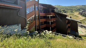 A modern multi-story building with a wooden exterior and large windows, surrounded by a grassy landscape and hills in the background. - 180m from shops, floor -1, view mountain, balcony, 60m², Méribel-Mottaret (Méribel - Les Allues)