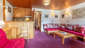 A cozy room with a wooden ceiling, a kitchen area with wooden cabinets, a red sofa with patterned cushions, and a wooden coffee table. - 180m from shops, floor -1, view mountain, balcony, 60m², Méribel-Mottaret (Méribel - Les Allues)
