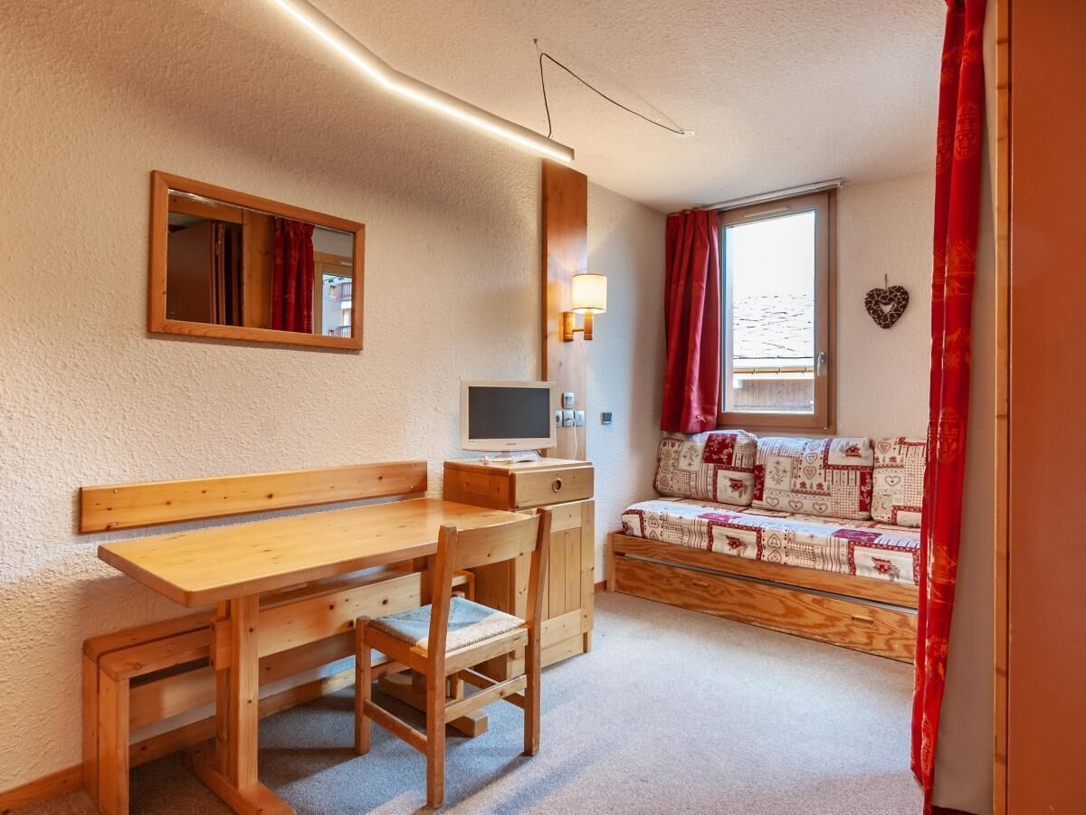 A cozy room with a wooden desk, chair, and bench, a small TV, and a window with red curtains.