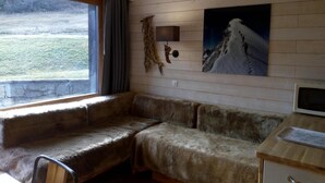 Et hyggeligt sommerhusinteriør med en bænk betrukket med pels, et billede af et bjerg på væggen og et træskrivebord med en stol.