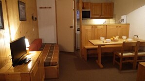 A compact room with a wooden table and bench, a television, and a kitchenette with wooden cabinets.