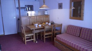 Una cocina compacta con armarios de madera, una mesa de comedor con sillas, un microondas y un tostador. El piso está cubierto por una alfombra morada y hay un sofá de madera con un cojín a cuadros.