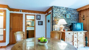 A dining area with a round table, chairs, a bowl of fruit, a stone wall, wooden cabinets, and a television. - 350m from ski slopes, balcony, tv, 40m², Méribel-Mottaret (Méribel - Les Allues)