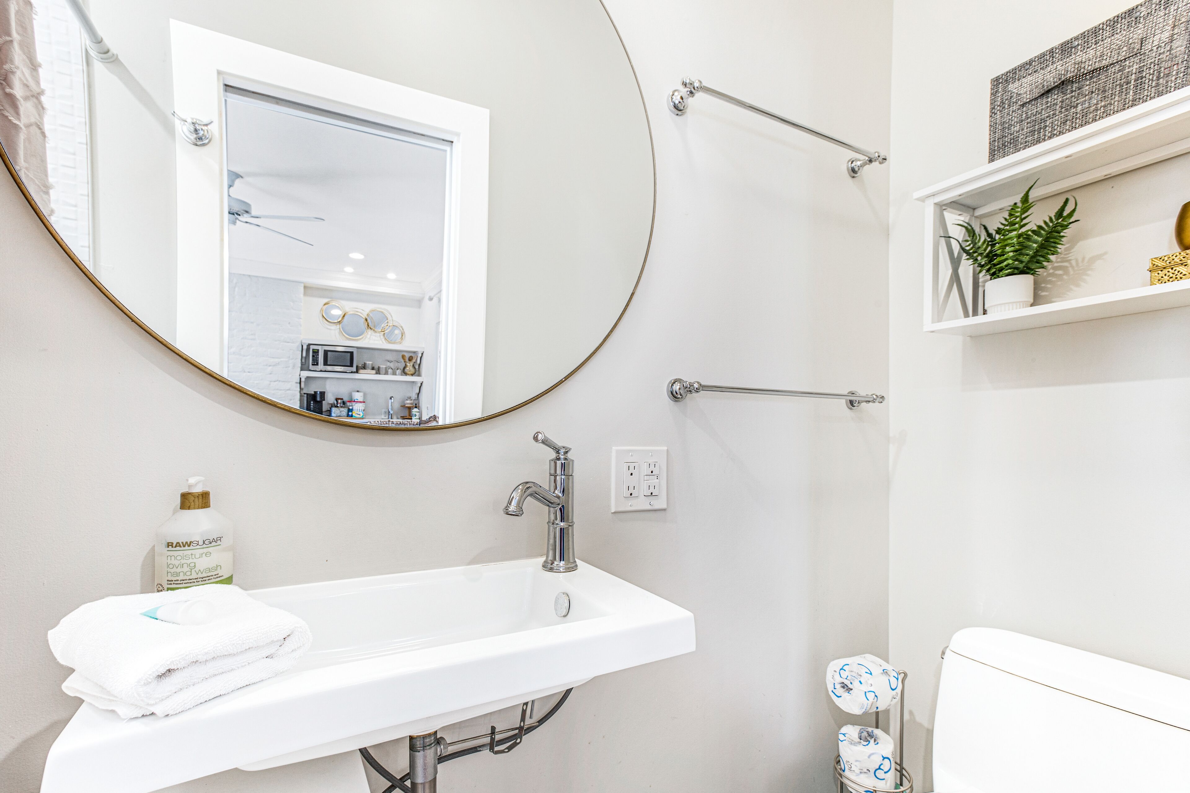 Une salle de bain moderne avec un lavabo blanc, un miroir rond, une serviette, une douchette à main et une étagère ornée d’une plante.