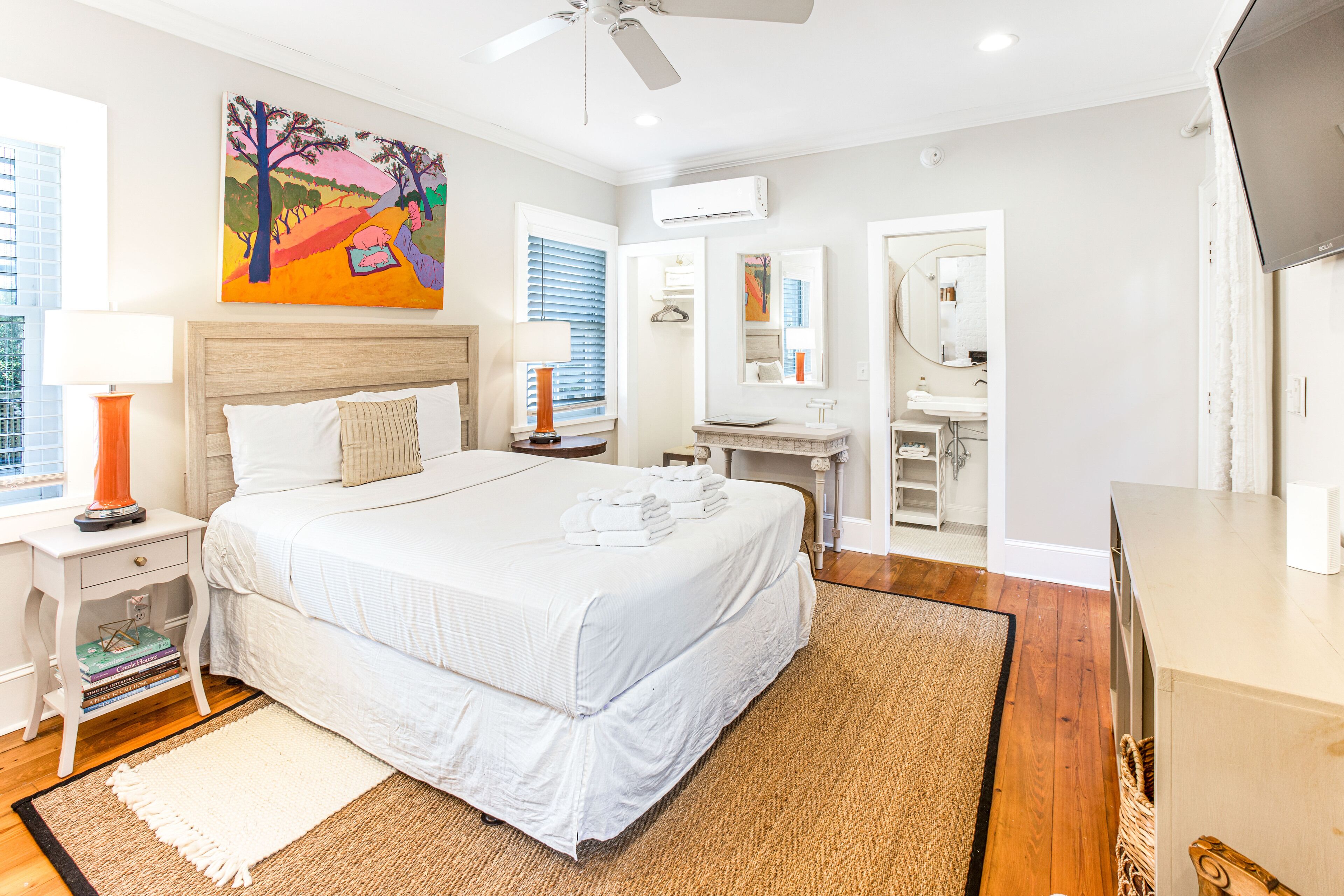 A bedroom with a large bed, bedside tables, a nightstand, a desk, a chair, a mirror, and an air conditioning unit.