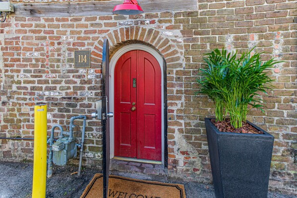 A red door with a number plate, a potted plant, and a yellow bollard.
