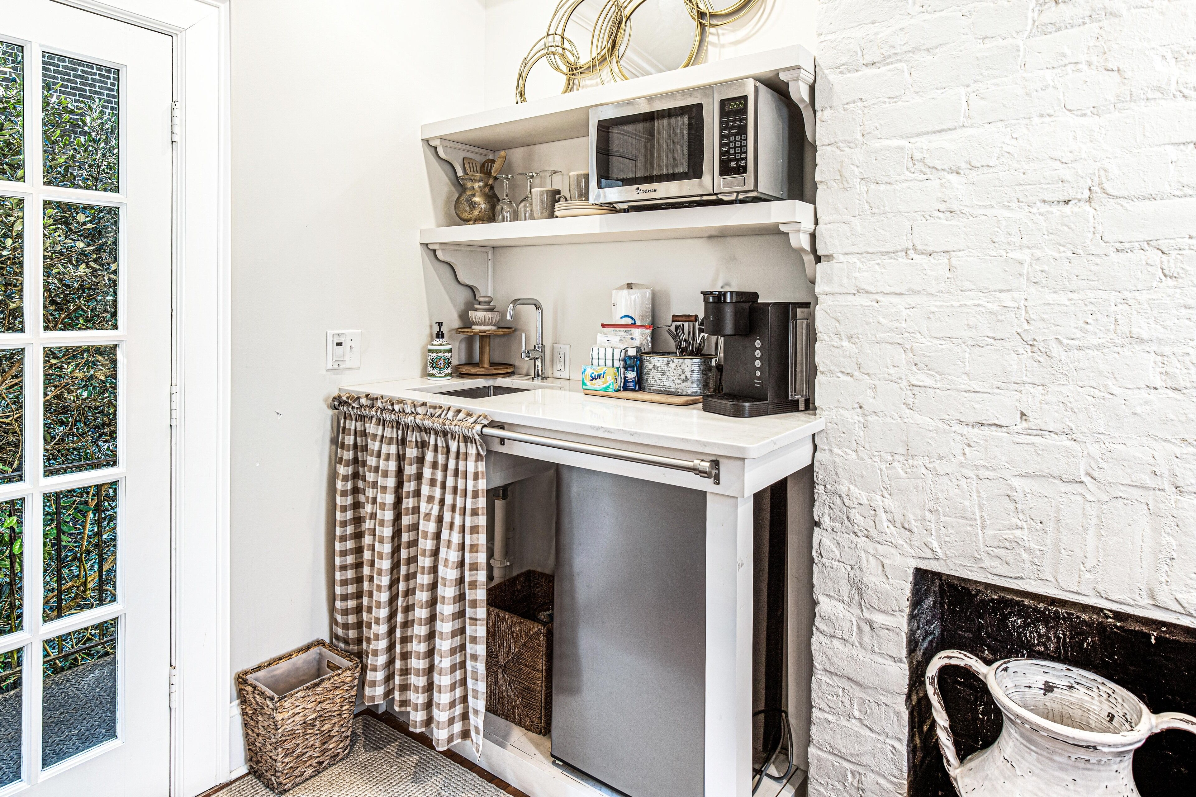 A compact kitchen with a microwave, coffee maker, and a small sink. There is a checkered towel hanging on the door, and a basket near the sink.