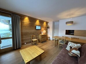 Uma sala de estar acolhedora com piso de madeira, um sofá, uma mesa de centro, uma TV na parede e uma área de jantar com mesa e cadeiras.