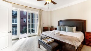 Un dormitorio con una cama grande, una mesita de noche, un banco y vistas a un edificio a través de un ventanal corredero.