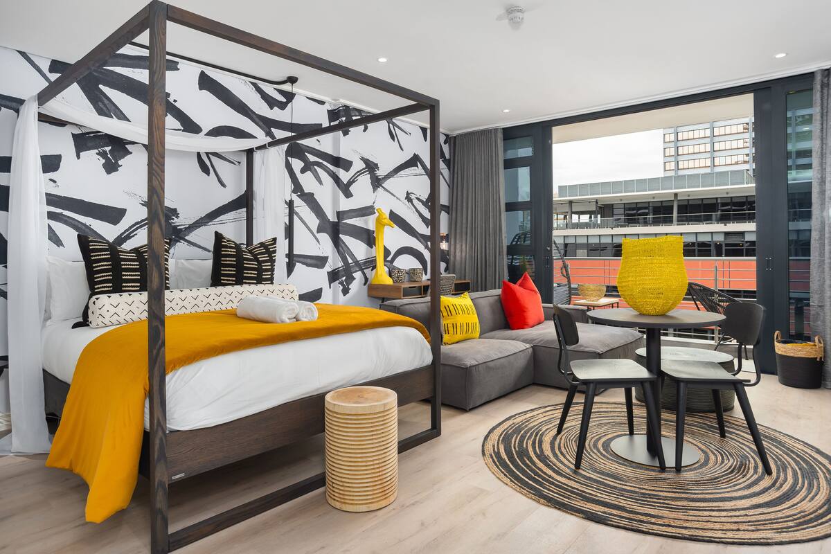 Une chambre d’hôtel moderne avec un grand lit, un canapé gris, une table à manger ronde et une vue sur un immeuble à travers de grandes fenêtres.
