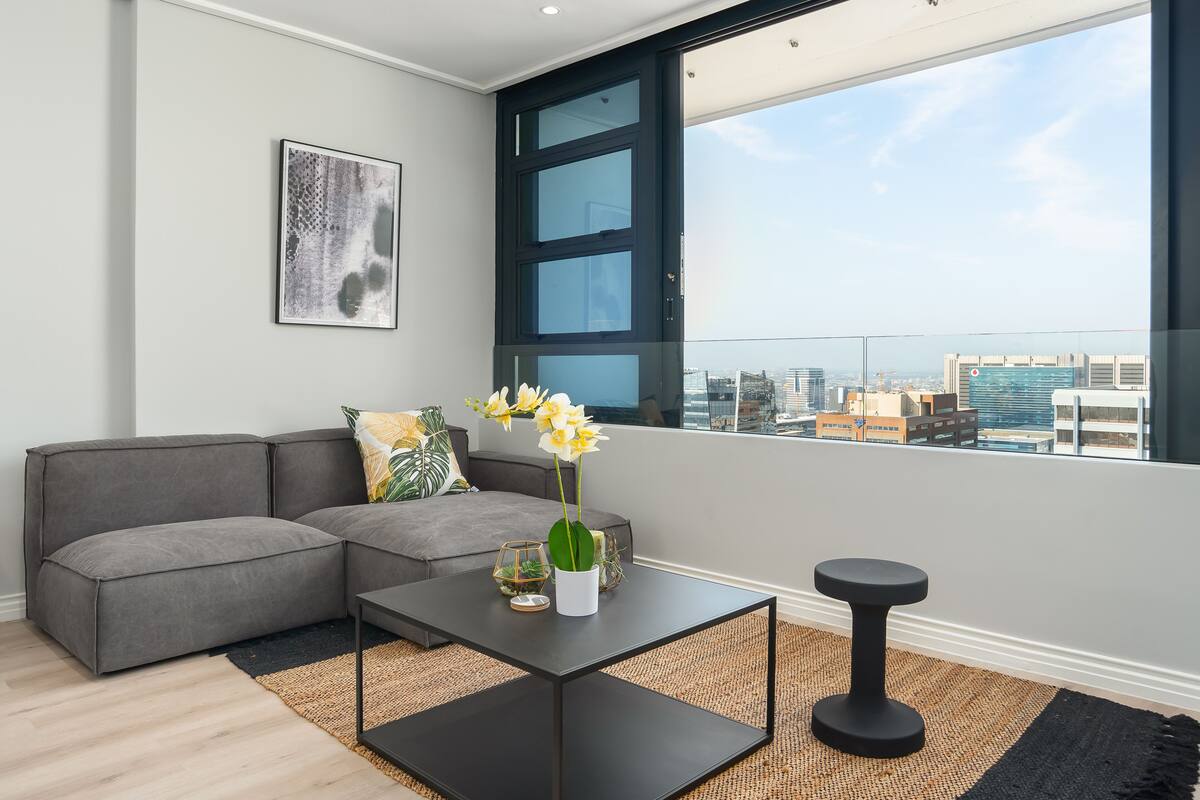 Un salon moderne avec un canapé d’angle, une table basse et une vue sur le paysage urbain grâce à une grande fenêtre.