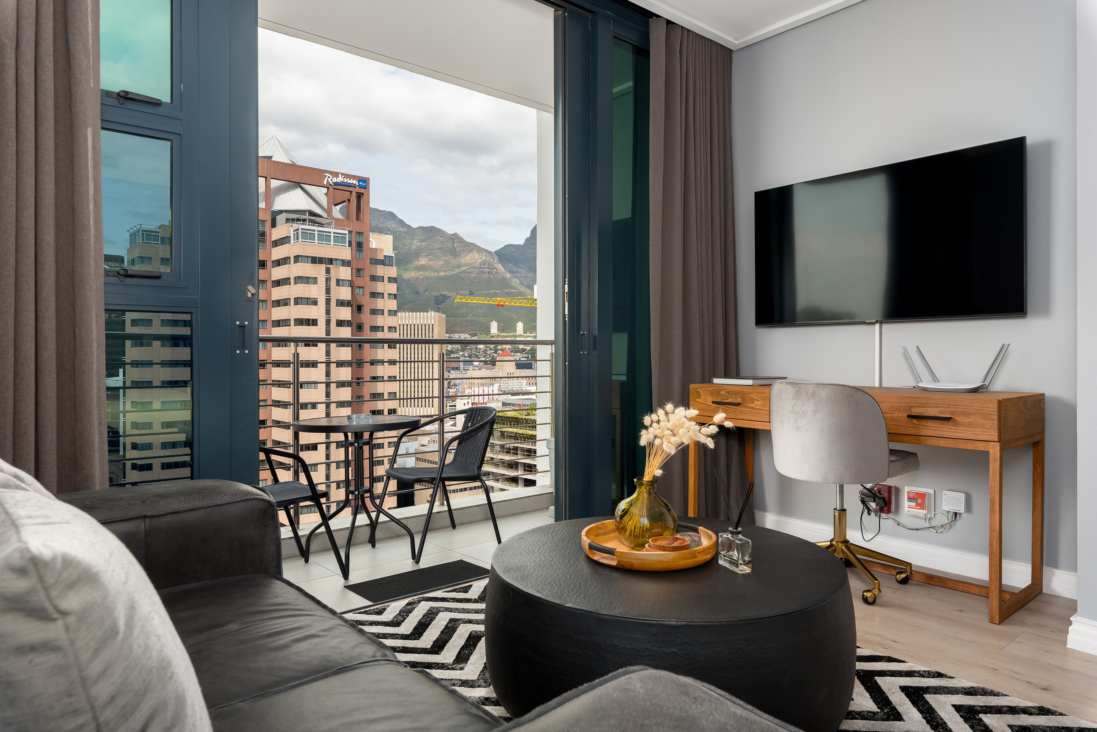 A modern living room with a sofa, a round coffee table, a TV, and a balcony with a city view.