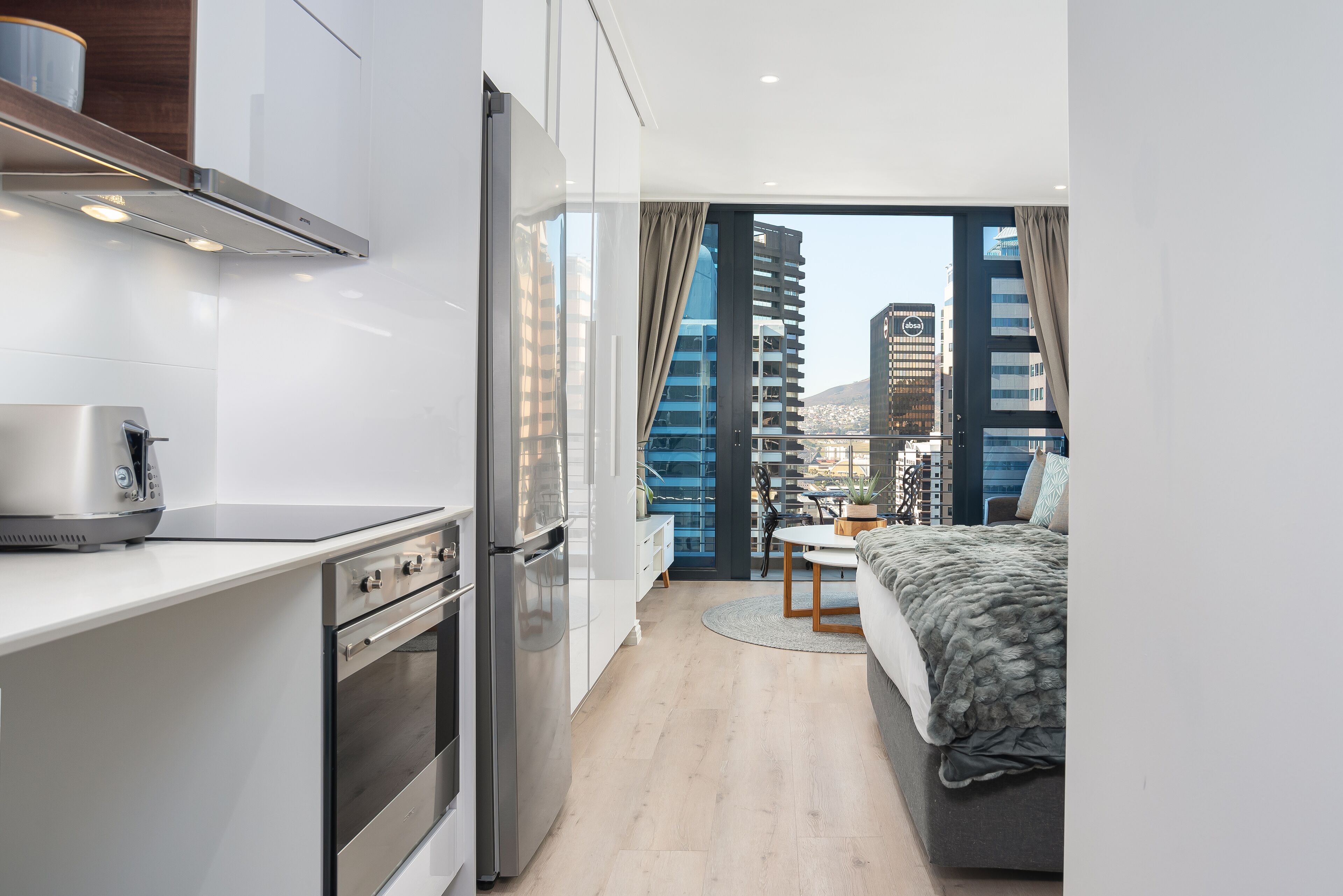 A modern hotel room with a kitchenette, a bed, a dining area, and a view of the cityscape.