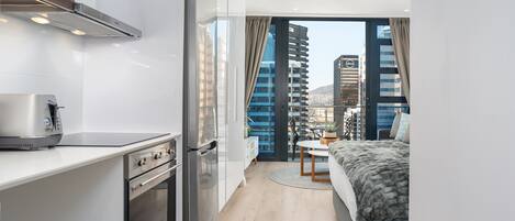 A modern hotel room with a kitchenette, a bed, a dining area, and a view of the cityscape.
