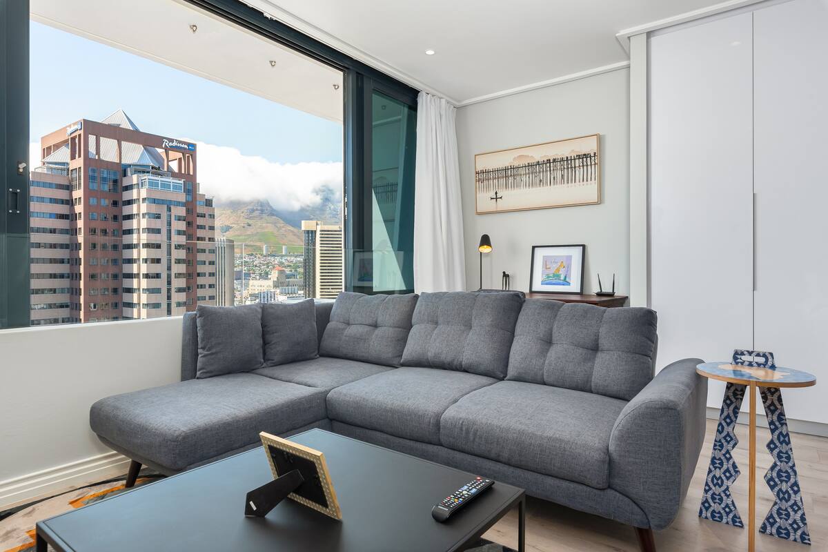 Un salon moderne avec un canapé d’angle gris, une petite table ronde et une grande fenêtre donnant sur la ville.