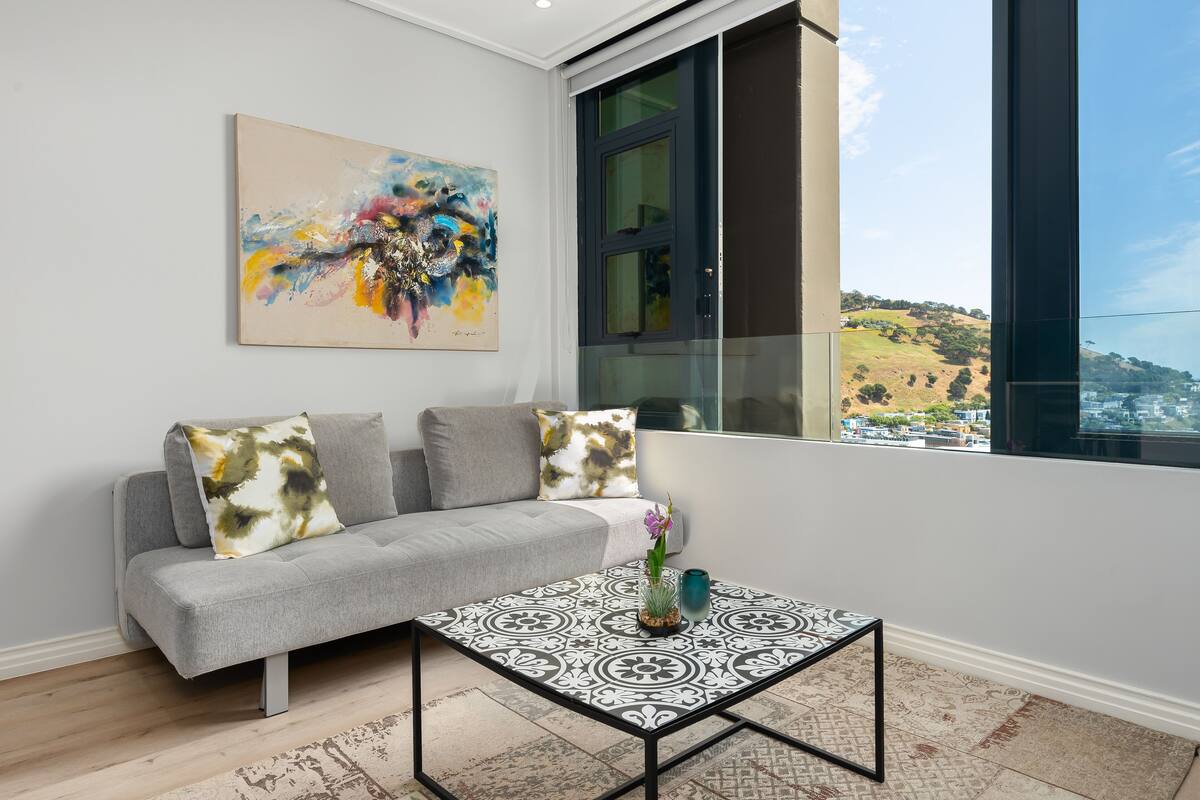 Un salon moderne avec un canapé gris, une table basse à motifs et une grande fenêtre donnant sur un paysage vallonné.