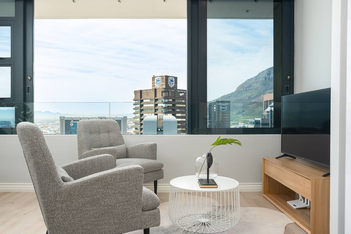 Un salon moderne avec des fauteuils gris à revêtement en tissu, une table basse ronde blanche, un meuble TV en bois et un grand téléviseur à écran plat.