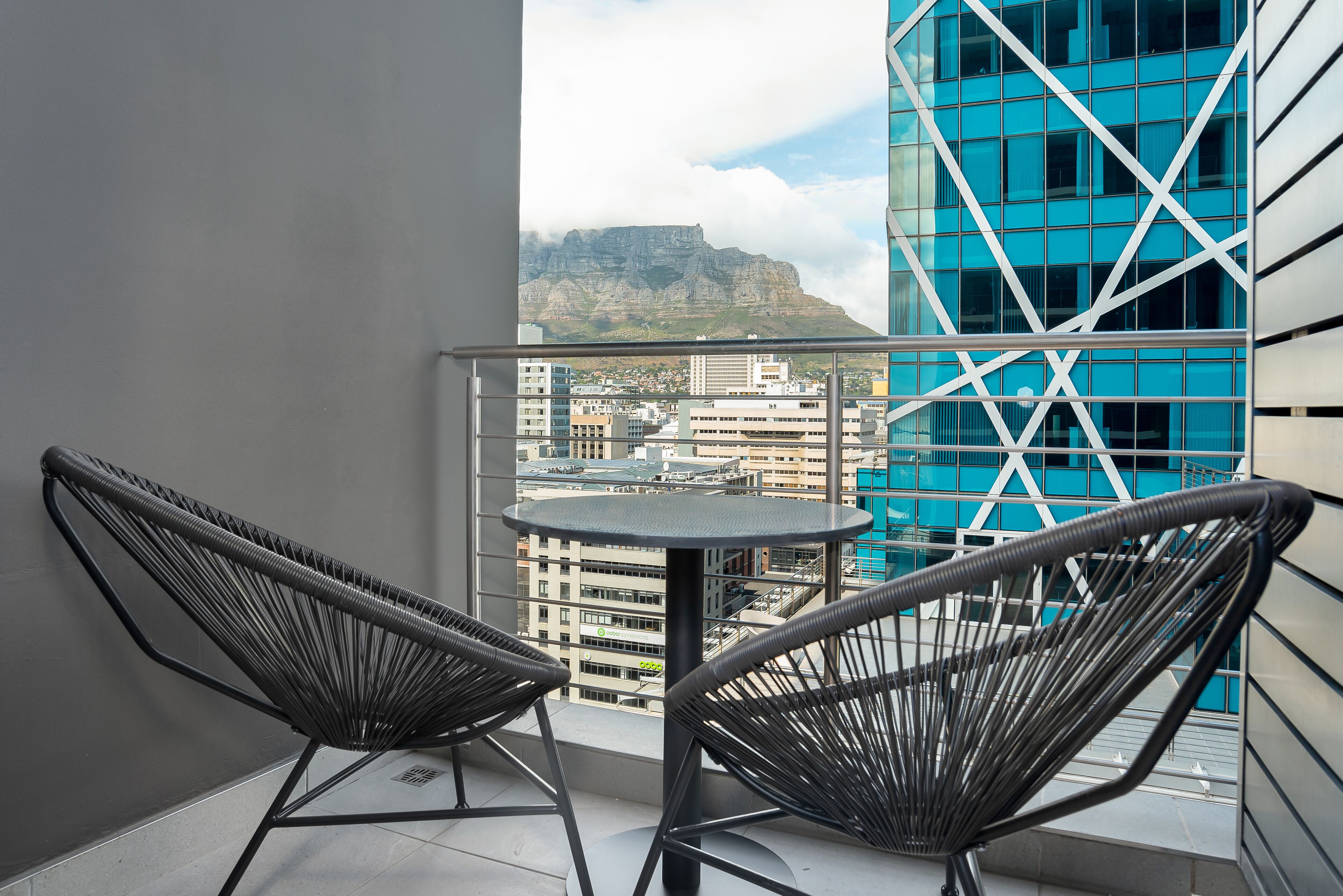 Un balcon avec deux fauteuils en osier et une petite table, d’où l’on peut admirer un paysage urbain avec un imposant bâtiment bleu et une montagne en arrière-plan.