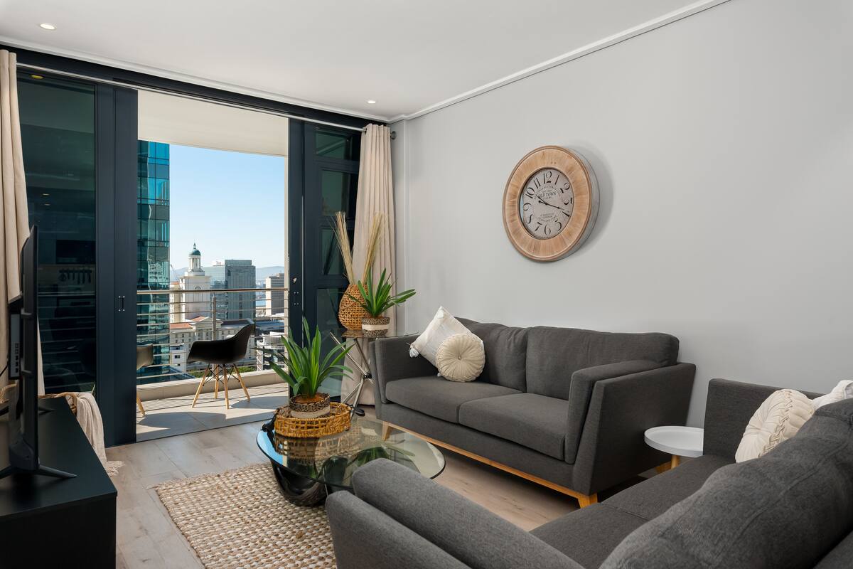 Un salon moderne avec un canapé gris, une horloge ronde en bois et une vue sur le paysage urbain, visible à travers une porte vitrée.