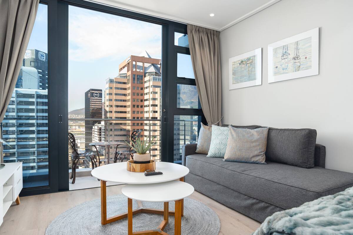 Un salon moderne avec un canapé gris, une petite table blanche et un balcon offrant une vue sur la ville.