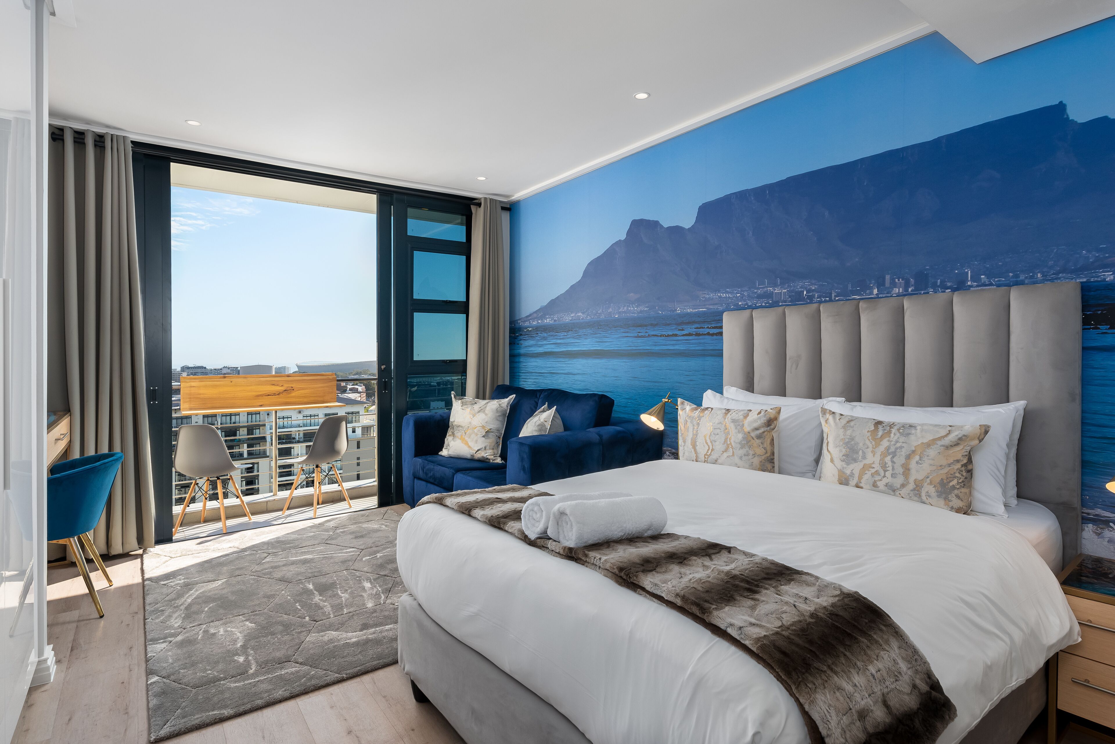 A modern hotel room with a large bed, a view of the city, and a balcony with seating.