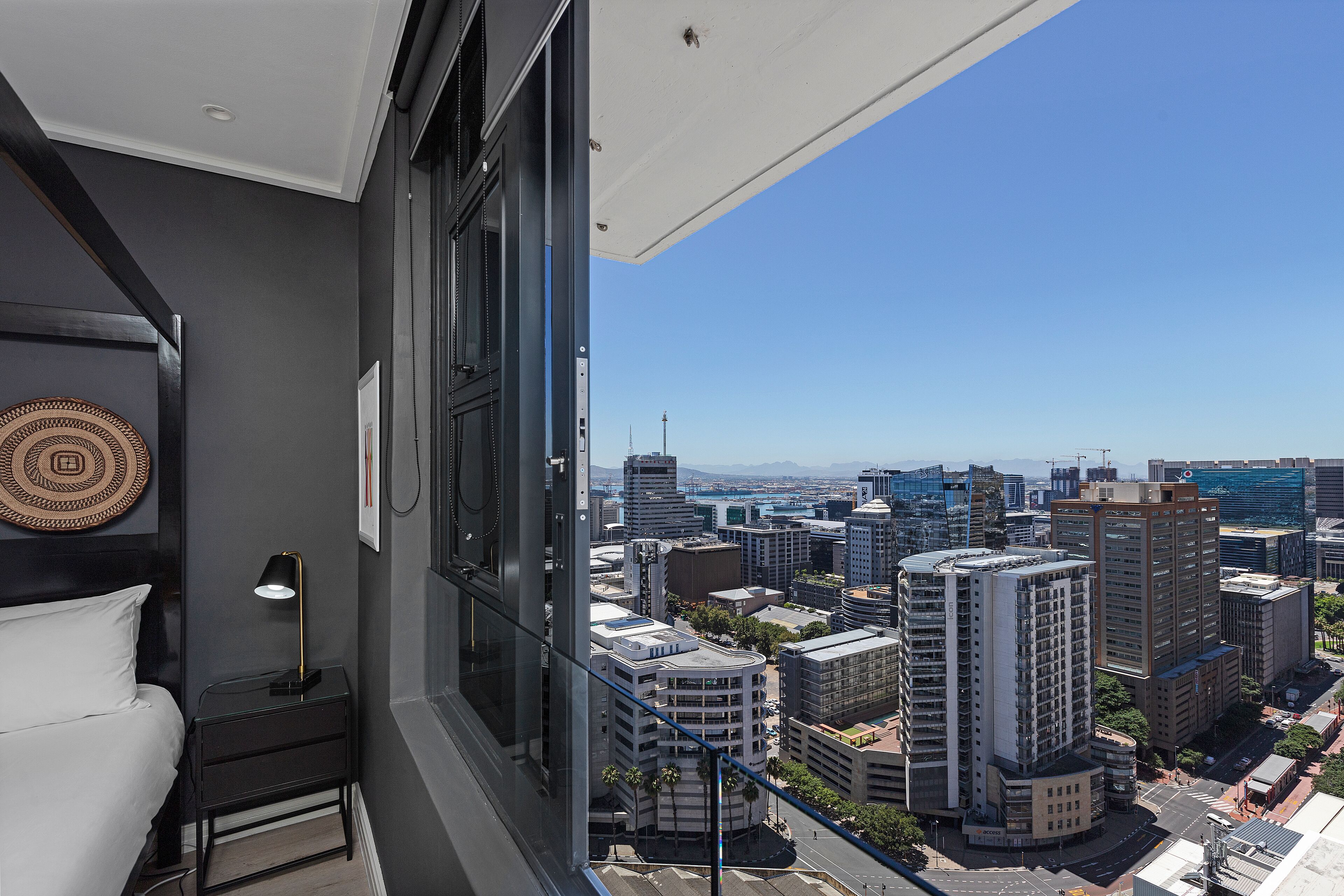 A bedroom with a city view, a bed, a nightstand, and a lamp.