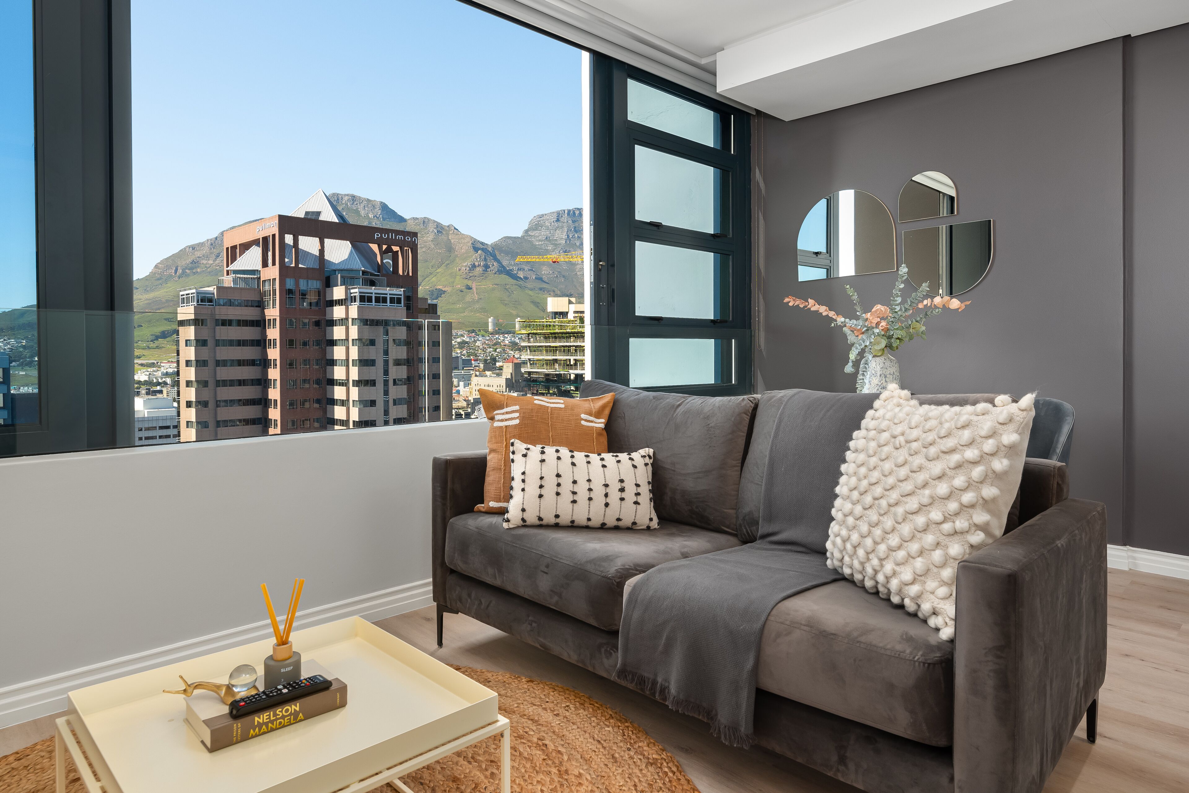 A modern living room with a grey sofa, a coffee table, and a view of a cityscape and mountains.