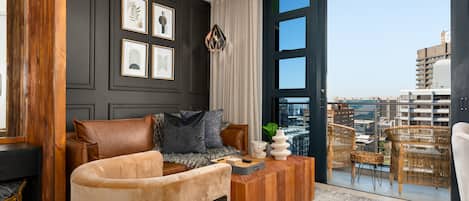 A modern living room with a sofa, coffee table, and wicker chairs. A large window offers a city view.