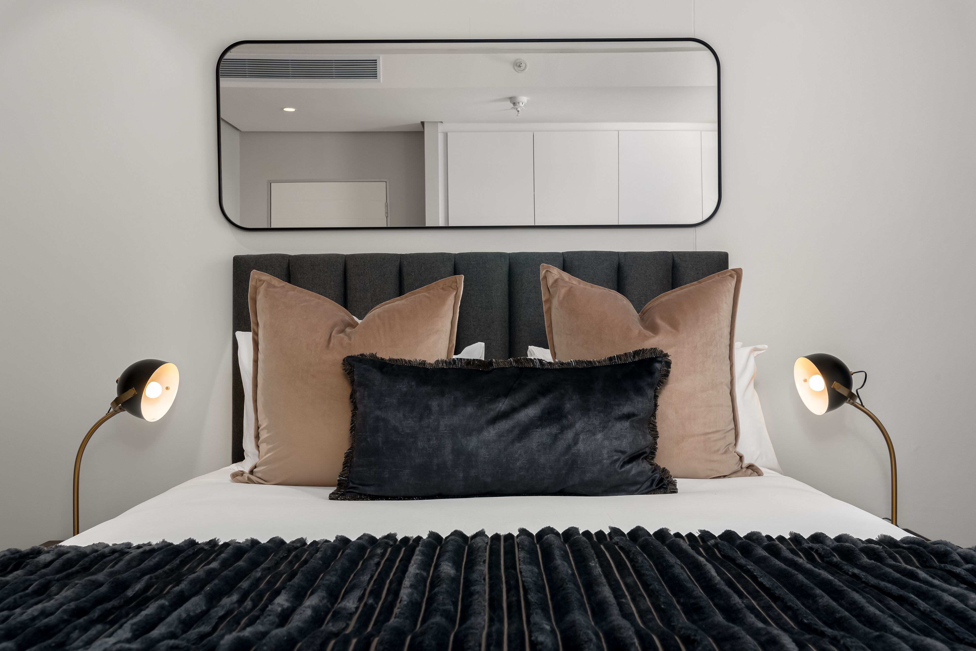 Cama bem arrumada com arranjo de almofadas preto e bege, cabeceira preta e duas luminárias de cabeceira.