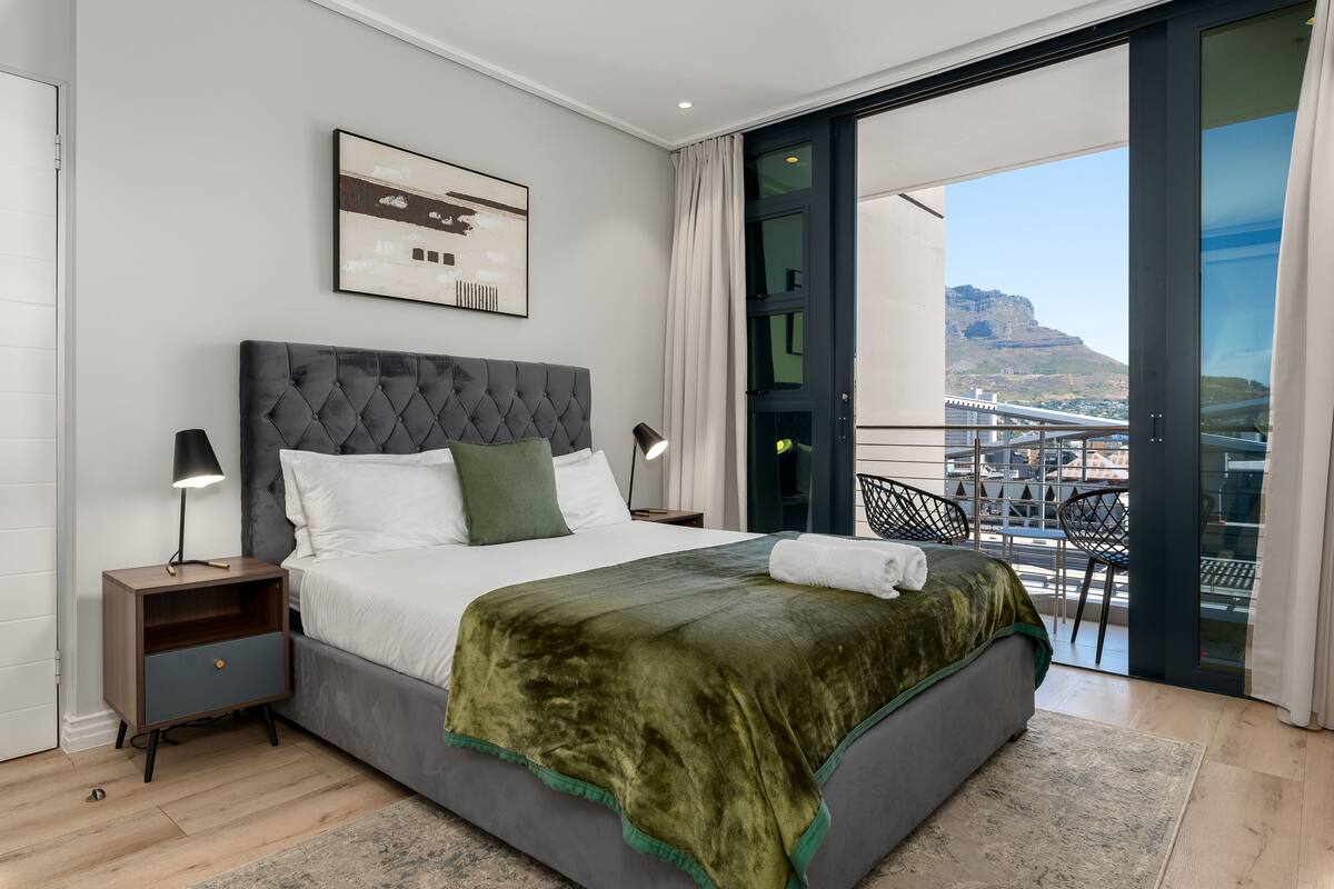Une chambre moderne avec un grand lit, une table de chevet, une lampe et une vue sur un balcon donnant sur un paysage urbain et des montagnes en arrière-plan.