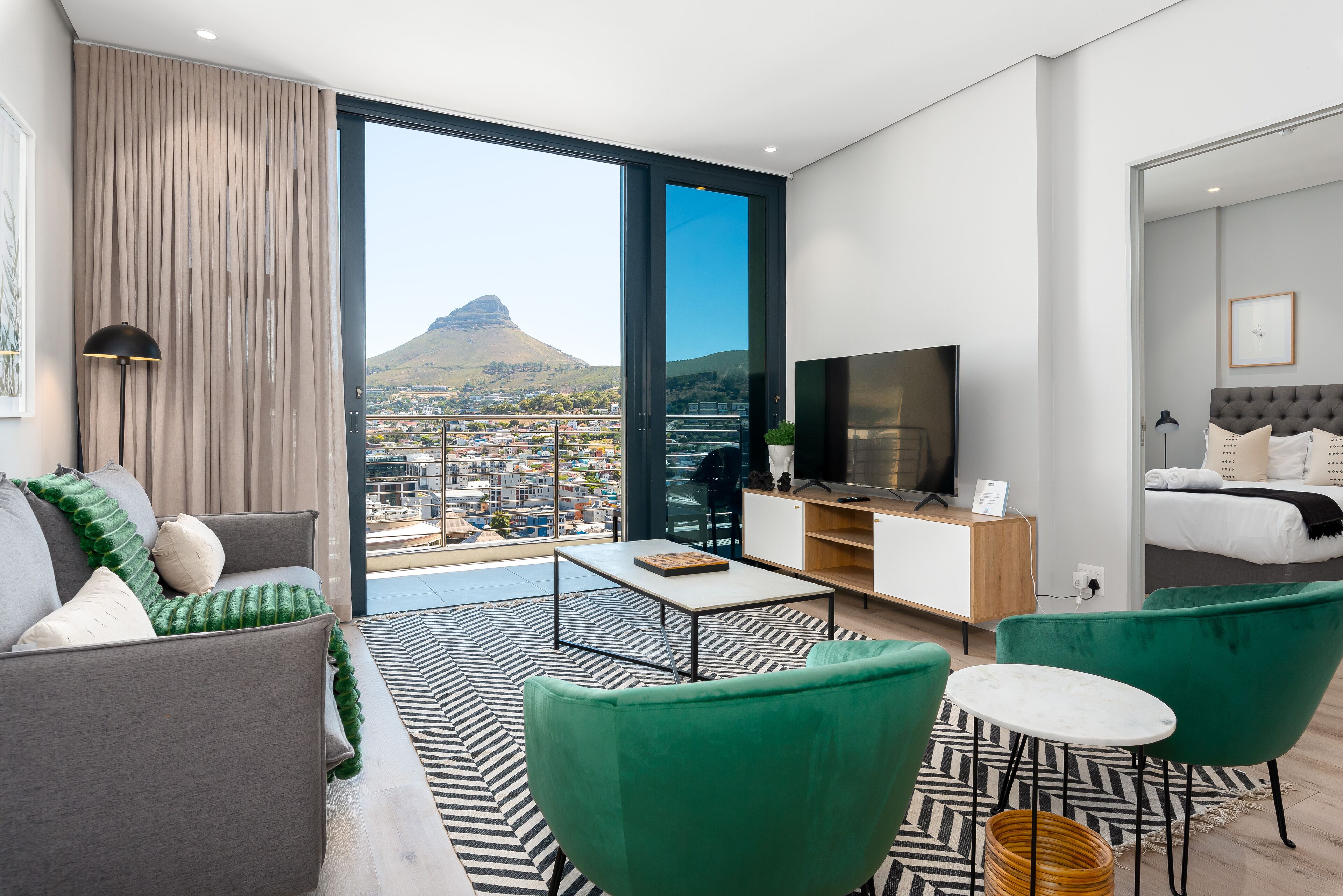 A modern hotel room with a large balcony, a sofa, a green armchair, a small table, a TV, and a bed.