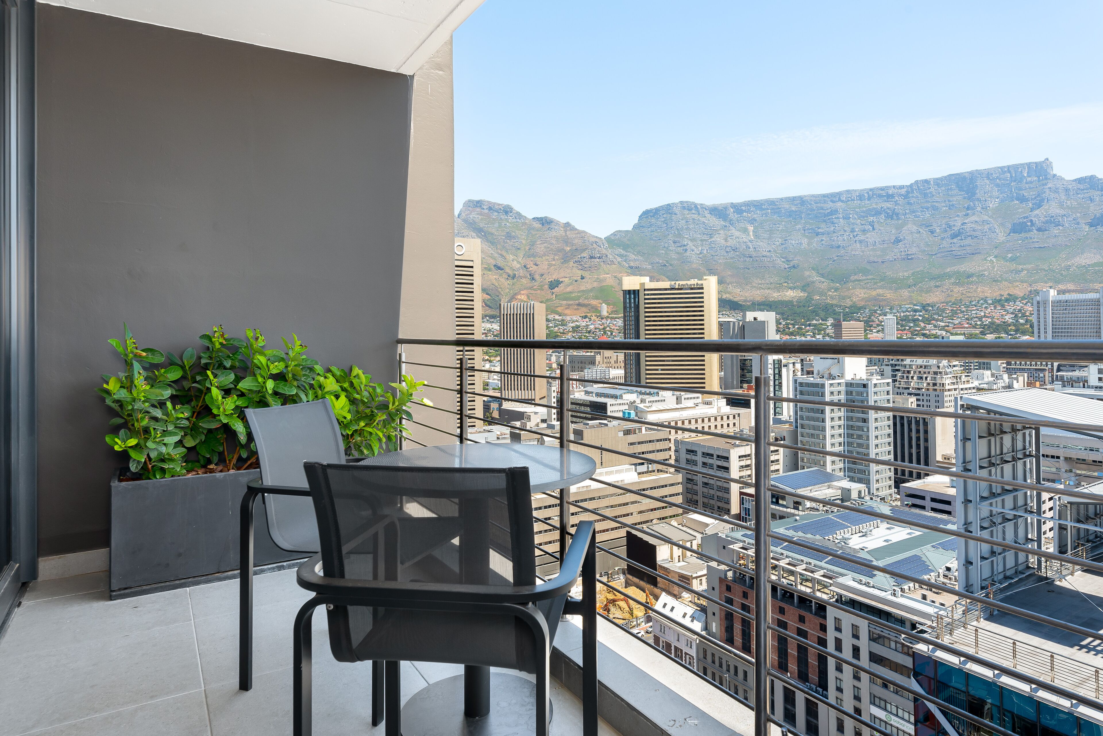Un balcón con mesa y sillas, una planta en maceta y vista a la ciudad con montañas al fondo.