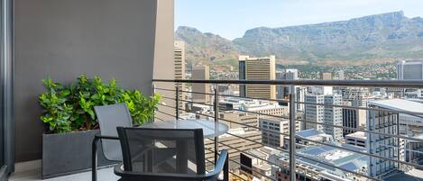 Un balcón con mesa y sillas, una planta en maceta y vista a la ciudad con montañas al fondo.