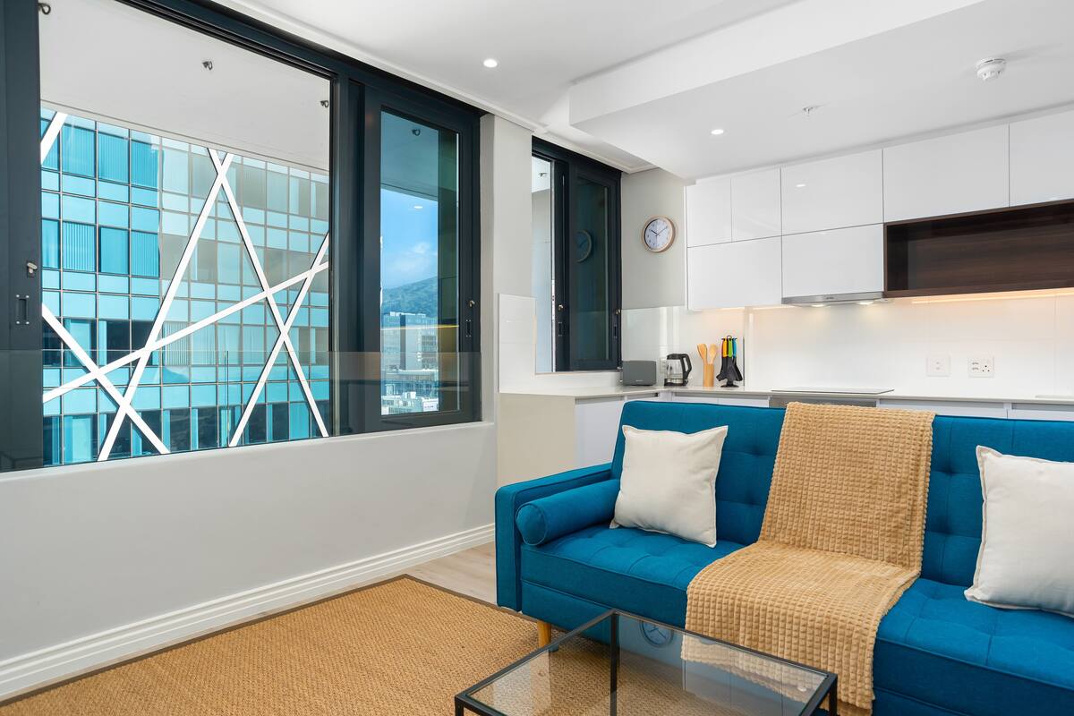 Un salon moderne avec un canapé bleu, une table basse en verre et une vue sur le paysage urbain, visible à travers de grandes fenêtres.