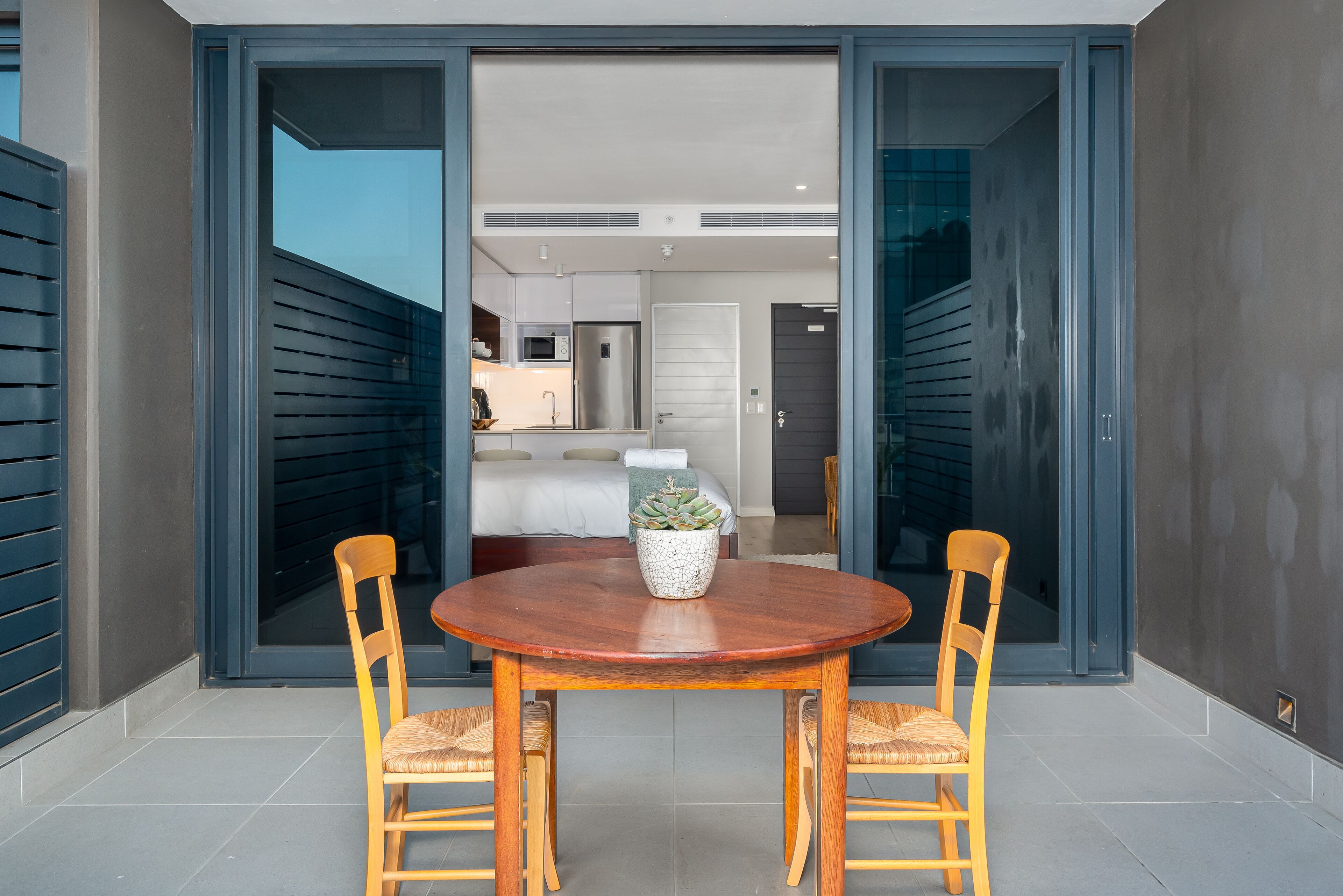 A modern dining area with a wooden table and chairs, a potted plant, and a view into a bedroom with a bed and a TV.