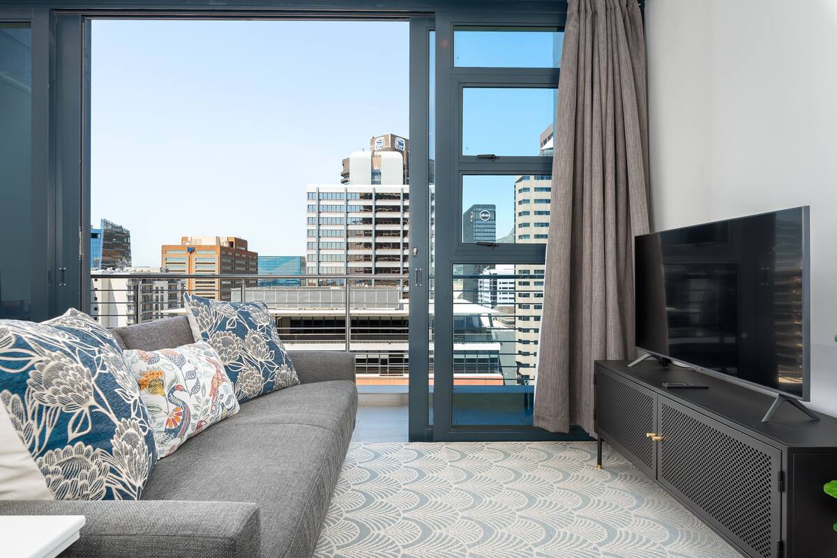 Un salon moderne avec un canapé gris, des coussins à motifs, un grand téléviseur à écran plat et une vue sur la ville, visible à travers une porte vitrée.