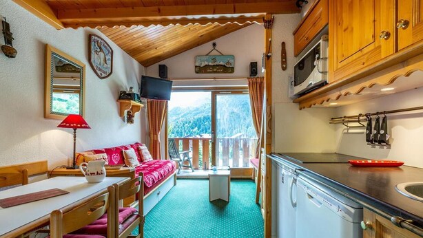 A cozy chalet-style room with a wooden interior, a dining area with a table and chairs, a kitchenette with a microwave and stove, and a large window offering a view of the mountains. - 90m from ski slopes, 3rd floor, view mountain, balcony, 27m², Méribel-Mottaret (Méribel - Les Allues)