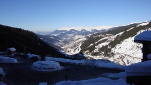 Un paysage enneigé avec une chaîne de montagnes en arrière-plan, une route qui traverse la vallée et un petit bâtiment sur la droite.