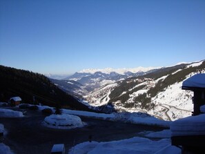 A snow-covered landscape with a mountain range in the background, a road leading through the valley, and a small building on the right.